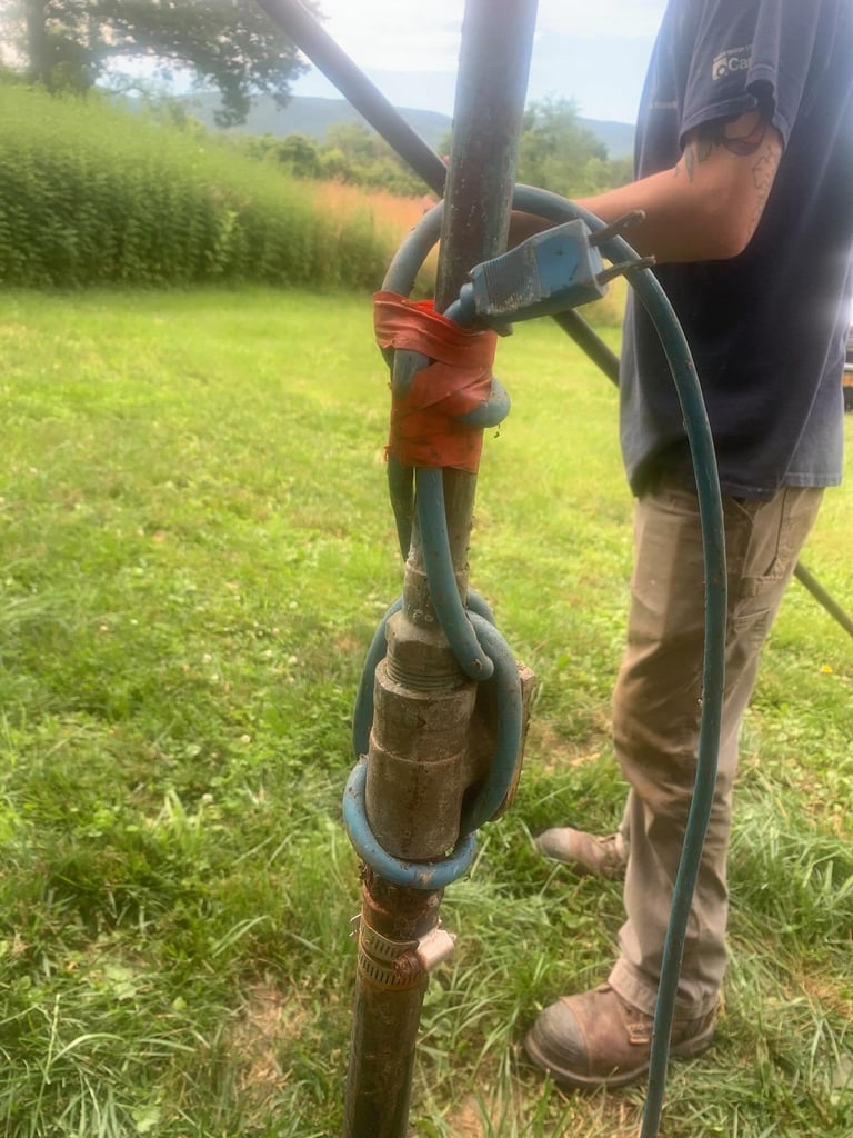 Person holding a water hose connected to a wooden post in a grassy field with mountains in background